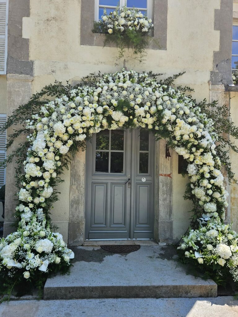 Grande arche florale entièrement blanche encadrant la porte d'un château : roses blanches, gypsophile, lisianthus et feuillage, avec deux compositions au sol