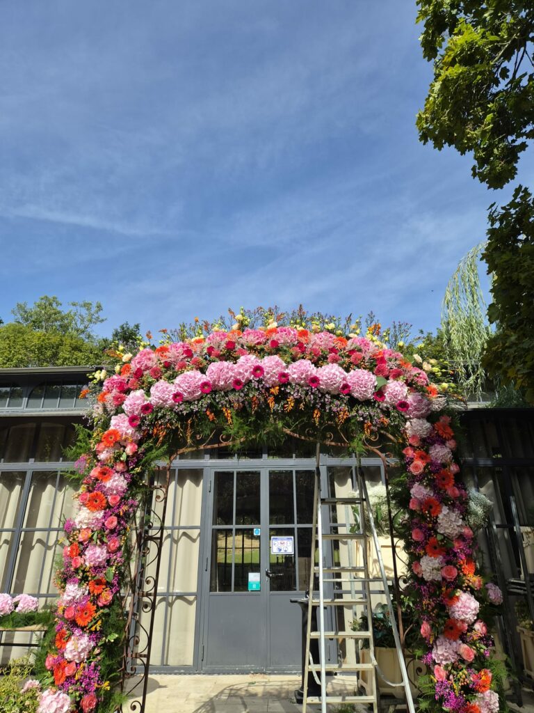 Grande arche florale de mariage montée devant une verrière : hortensias roses, gerberas orange et fuchsia, roses et feuillage, sur une structure en fer forgé