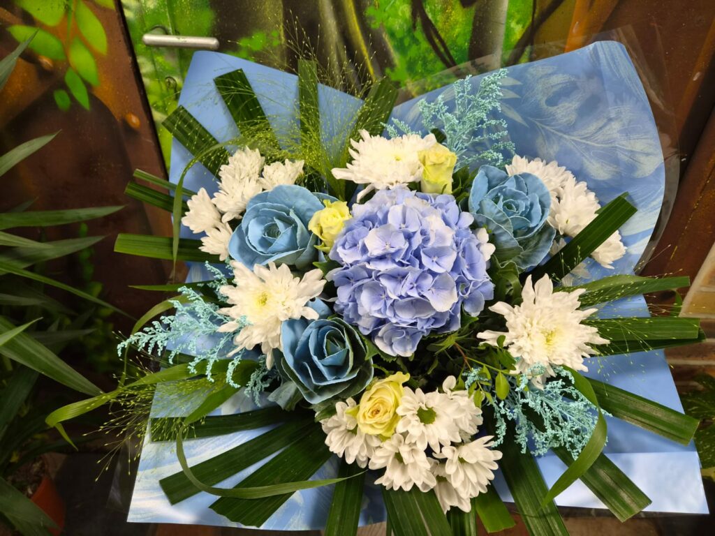 Bouquet rond bleu et blanc : hortensia bleu au centre, roses teintées bleu, chrysanthèmes blancs et petites roses jaune pâle, dans un papier bleu