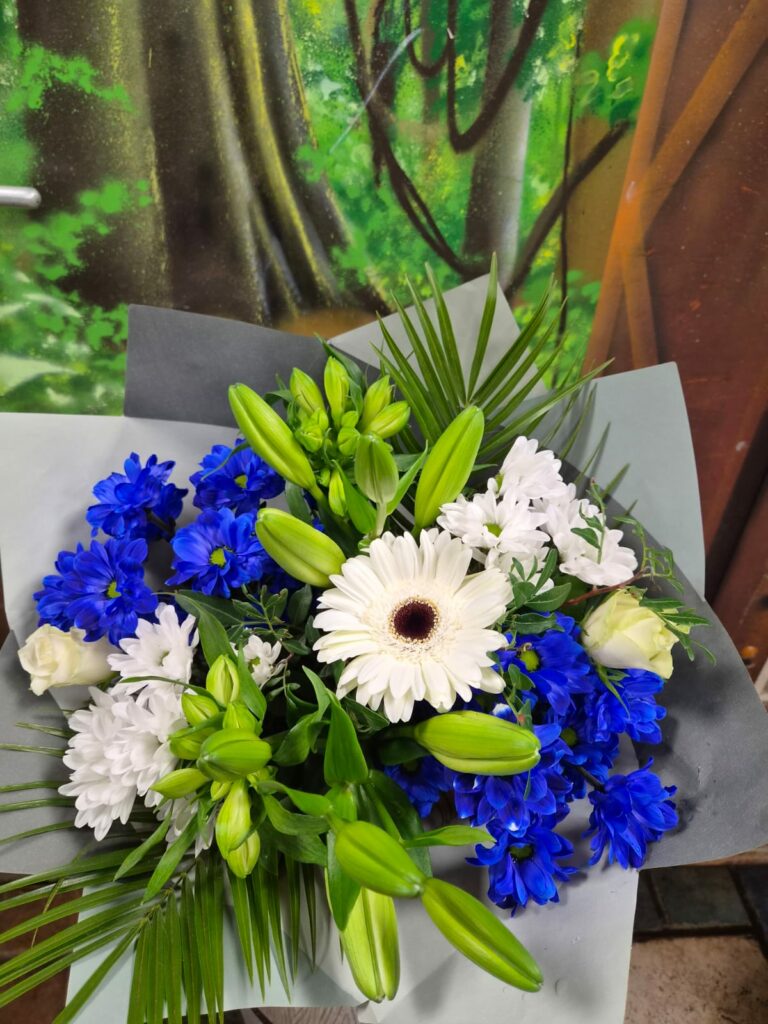 Bouquet bleu et blanc : gerbera blanc au centre, chrysanthèmes bleu vif et blancs, roses blanches et nombreux boutons de lis, dans un papier gris