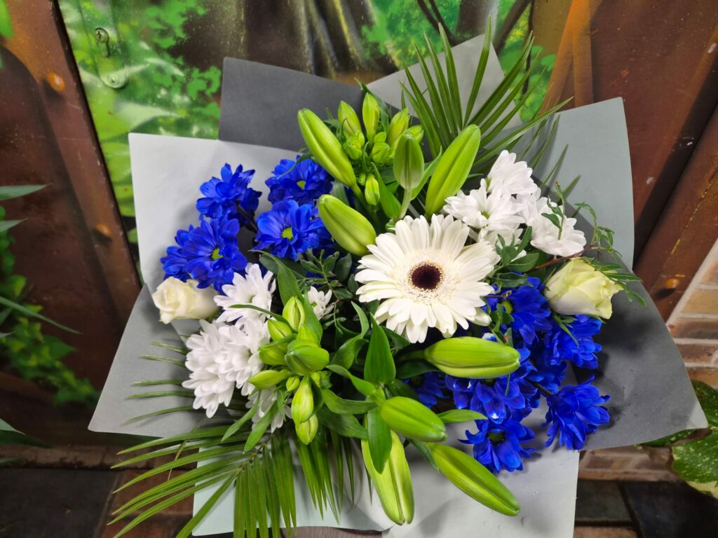 Bouquet bleu roi et blanc : gerbera blanc, chrysanthèmes bleu roi et blancs, roses blanches et boutons de lis, dans un papier gris