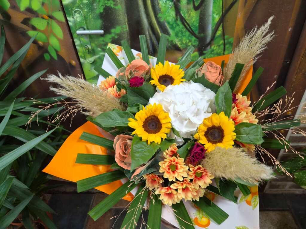 Bouquet aux tons chauds : tournesols, hortensia blanc, roses cuivrées, chrysanthèmes orangés et plumes de pampa séchées, dans un papier orange