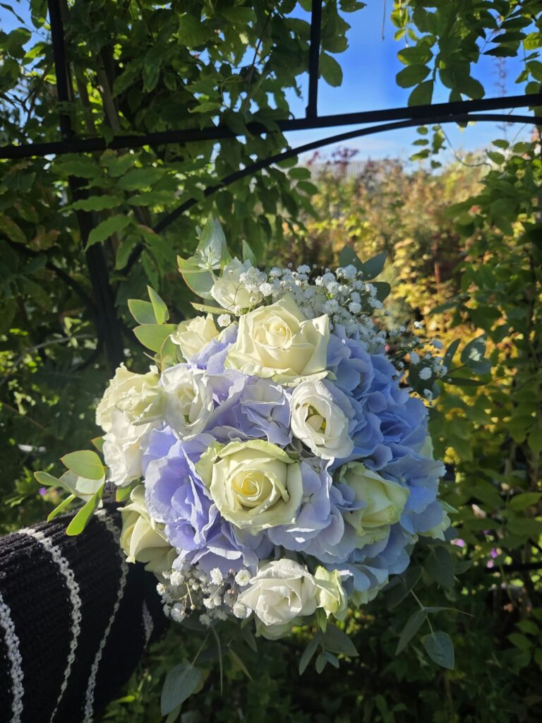 Bouquet de mariée rond bleu et blanc : hortensia bleu lavande, roses blanches, lisianthus et gypsophile, sur fond de jardin