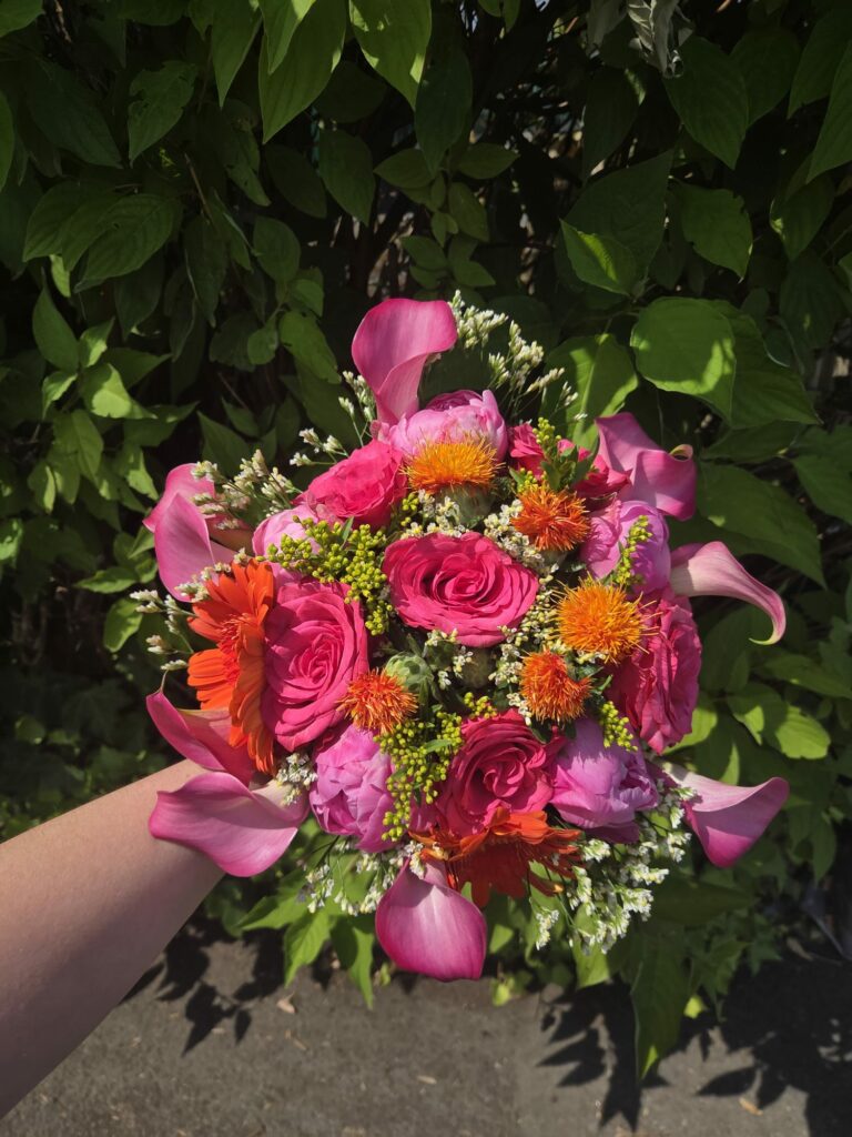 Bouquet de mariée aux tons fuchsia tenu à la main : roses fuchsia, arums roses, pivoines, carthame orange, gerbera et solidago