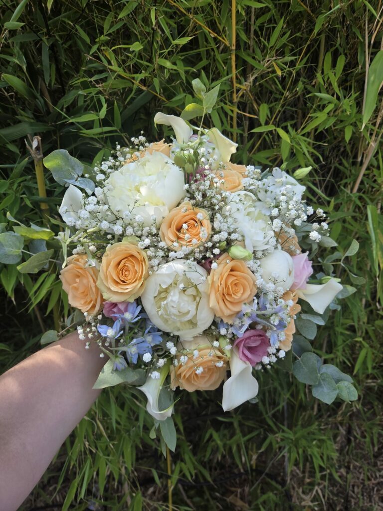 Bouquet de mariée rond tenu à la main : pivoines blanches, roses abricot, arums blancs, gypsophile, delphinium bleu pâle et eucalyptus