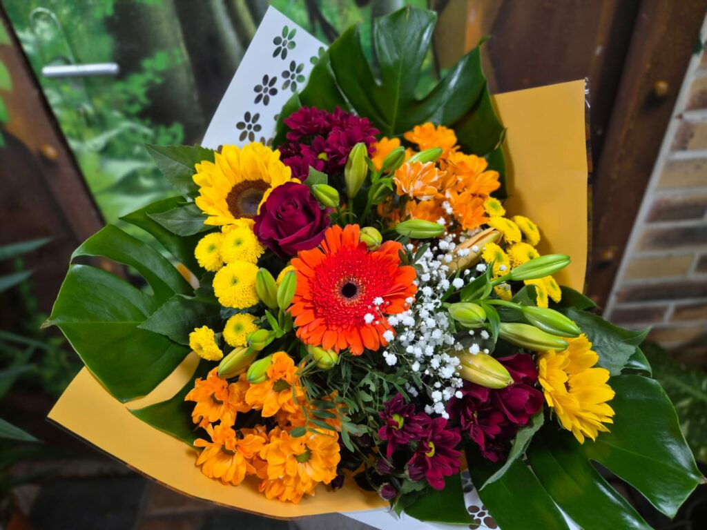 Bouquet multicolore aux tons chauds : tournesols, gerbera orange, roses pourpres, chrysanthèmes orange, jaunes et bordeaux, gypsophile et feuilles de monstera