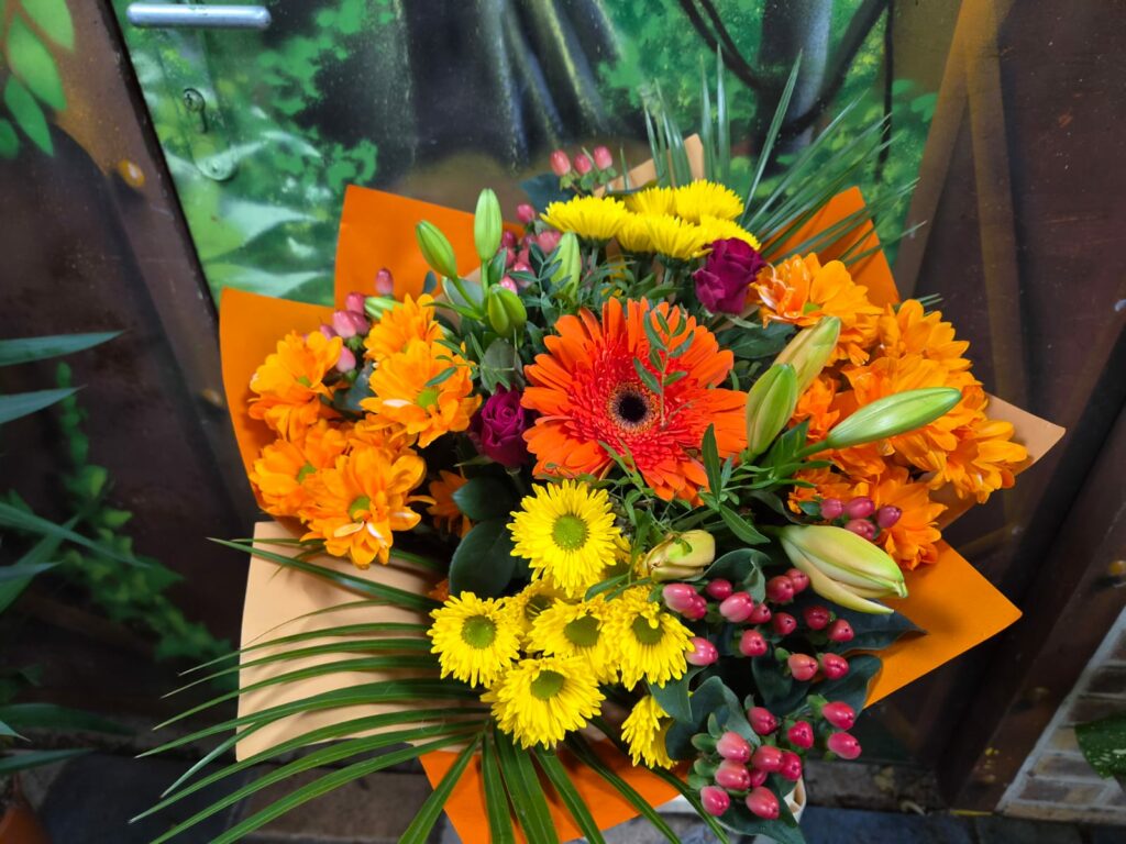 Bouquet aux tons orange et jaune : gerbera orange, chrysanthèmes orange et jaunes, petites roses pourpres, boutons de lis et baies d'hypericum, dans un papier orange