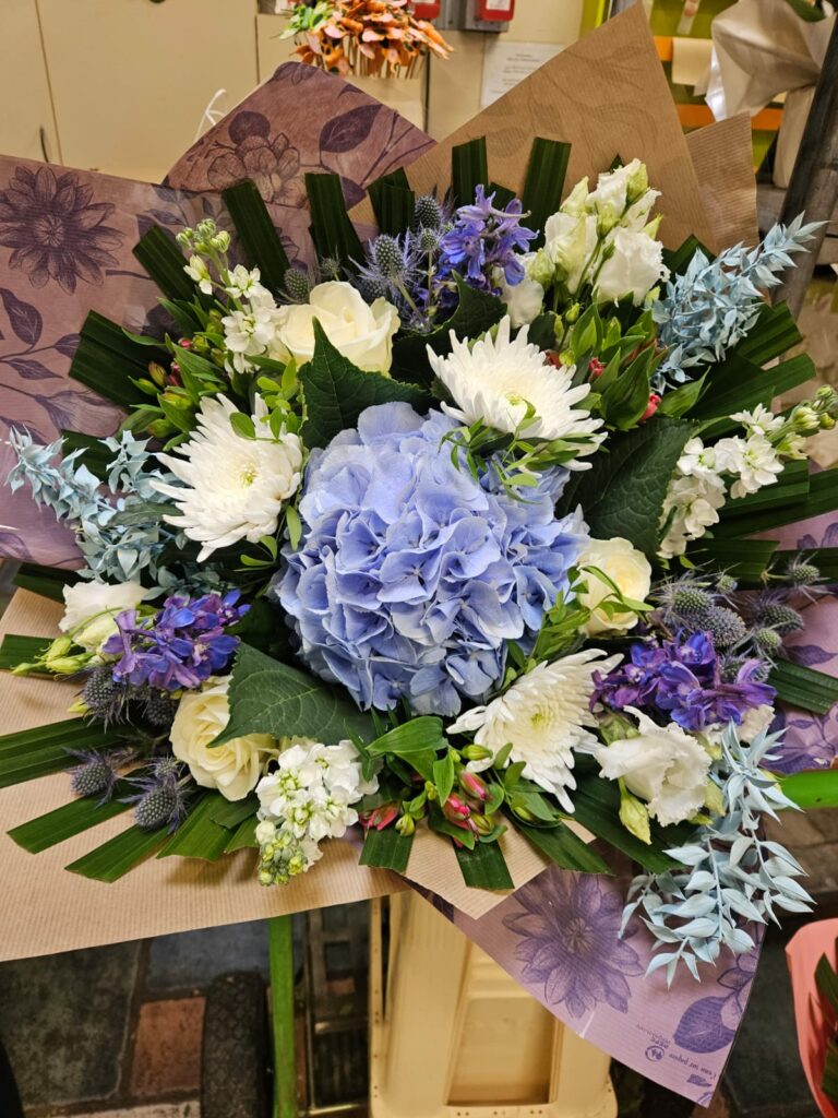 Bouquet rond bleu et blanc : hortensia bleu au centre, delphiniums violets, chardons bleus, roses et chrysanthèmes blancs, dans un papier mauve à motifs floraux