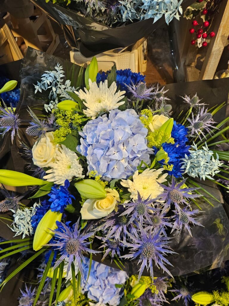 Bouquet rond aux tons bleus : hortensia bleu pâle, chardons bleus, chrysanthèmes blancs, roses blanches et boutons de lis, emballé dans un papier noir