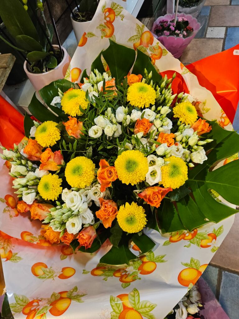 Bouquet rond aux tons jaunes et orange : chrysanthèmes pompons jaunes, roses orange, lisianthus blancs et grandes feuilles de monstera