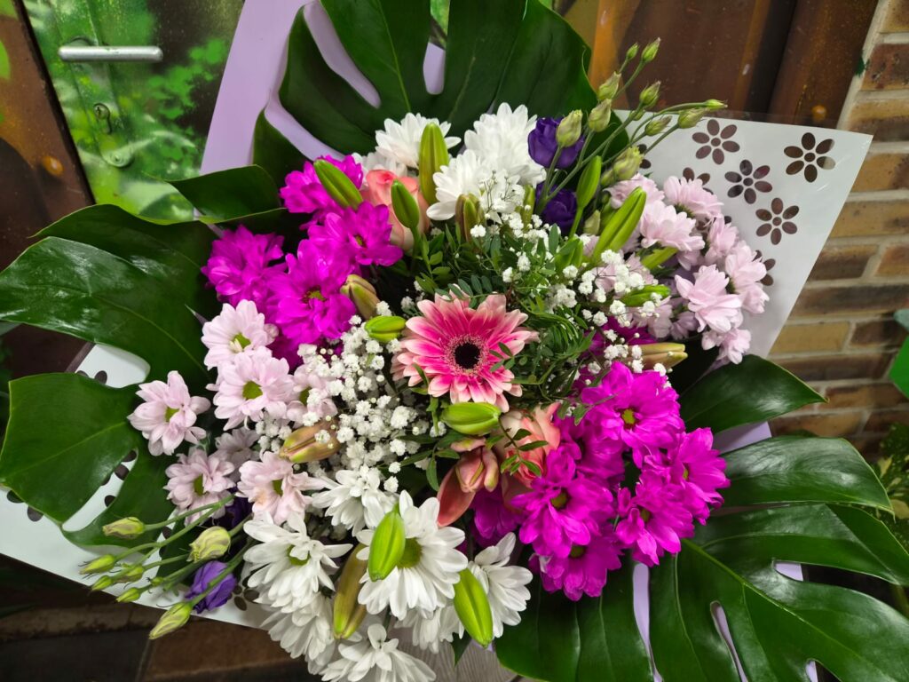 Bouquet aux tons roses : gerbera rose, chrysanthèmes fuchsia, rose pâle et blancs, lisianthus violets et gypsophile, sur de larges feuilles de monstera