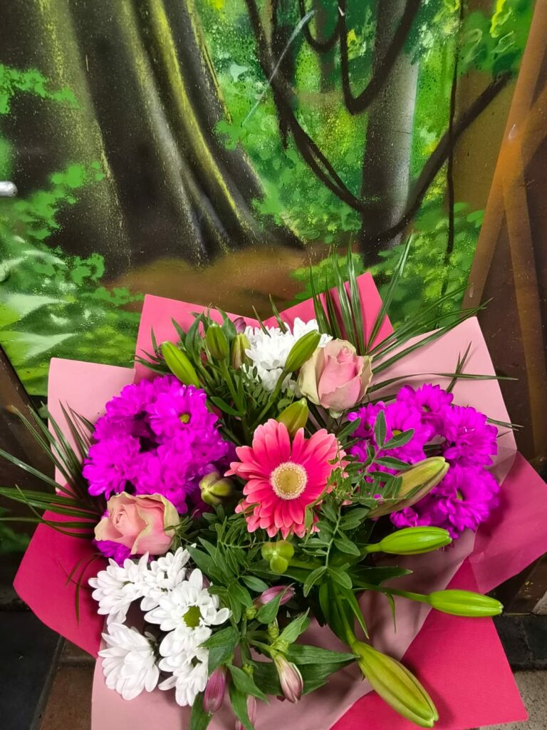 Bouquet aux tons roses et fuchsia : gerbera rose, chrysanthèmes fuchsia et blancs, roses pâles et boutons de lis, dans un papier rose vif
