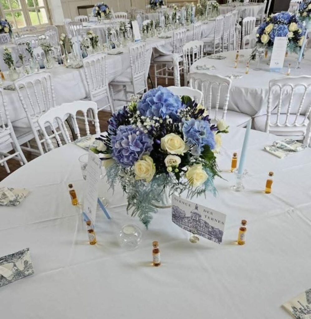 Centre de table de mariage dans une salle de réception : hortensias bleus, roses crème, lisianthus et gypsophile, sur des tables rondes nappées de blanc