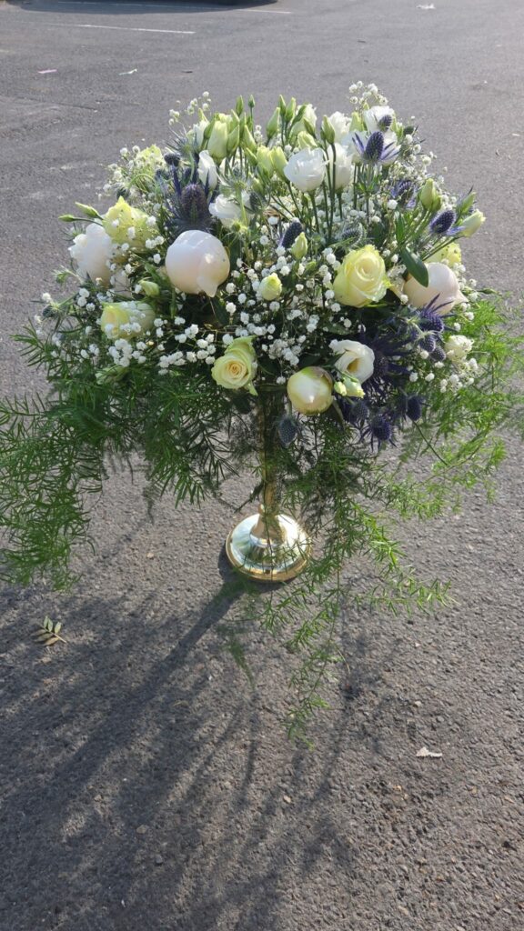 Centre de table de mariage monté sur un pied doré : pivoines blanches, roses ivoire, lisianthus, chardons bleus, gypsophile et feuillage d'asparagus retombant