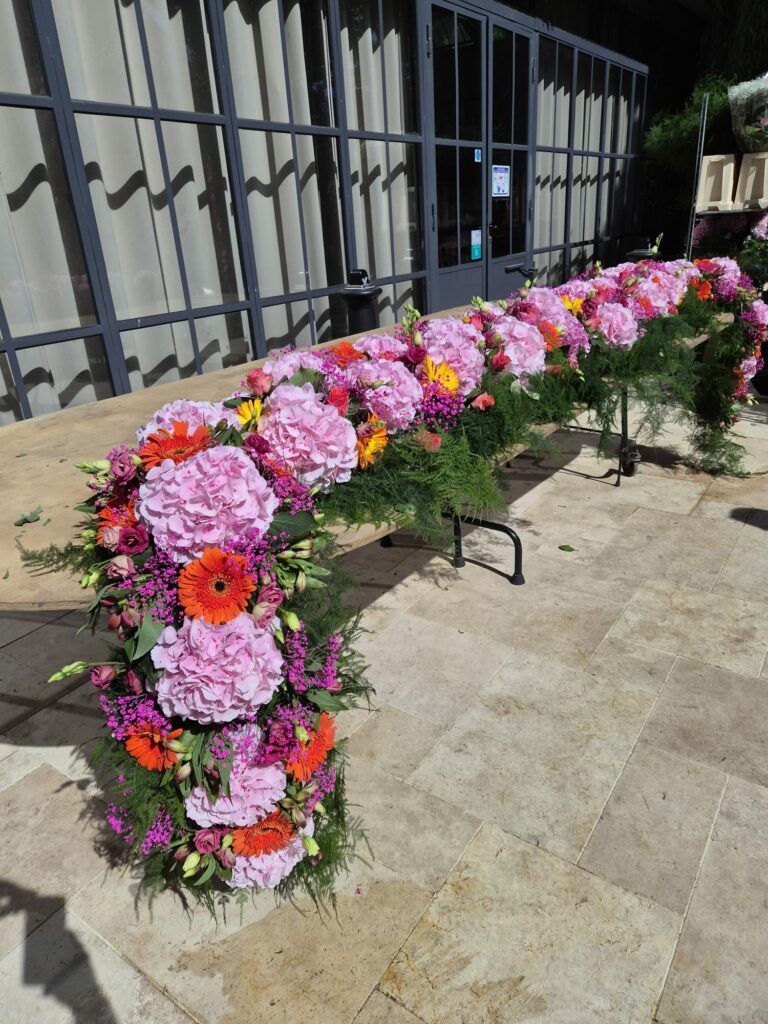 Chemin de table fleuri pour un mariage : hortensias roses, gerberas orange, lisianthus et feuillage d'asparagus, disposé sur une longue table en bois