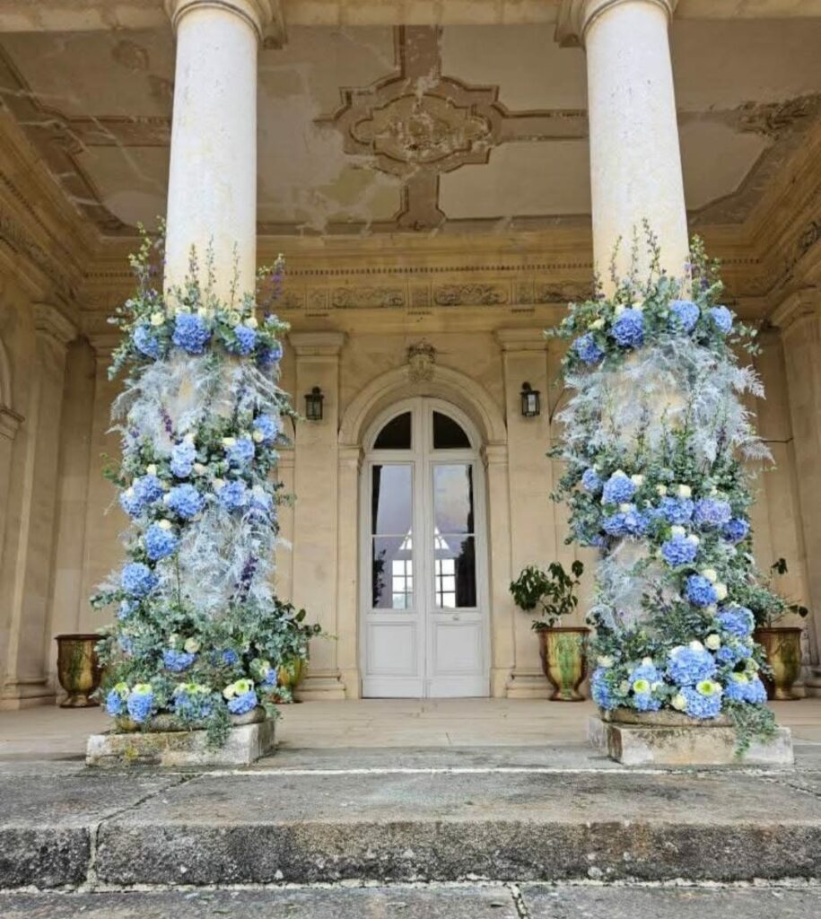 Deux colonnes de château habillées de fleurs pour un mariage : hortensias bleus, roses blanches, delphiniums et pampa blanchie, devant une porte à fronton