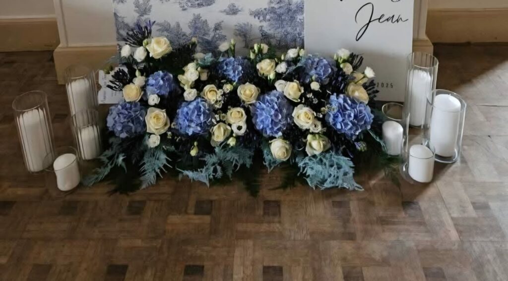 Longue composition florale posée au sol devant la table d'honneur : hortensias bleus, roses ivoire, lisianthus et feuillage bleuté, entourée de bougies