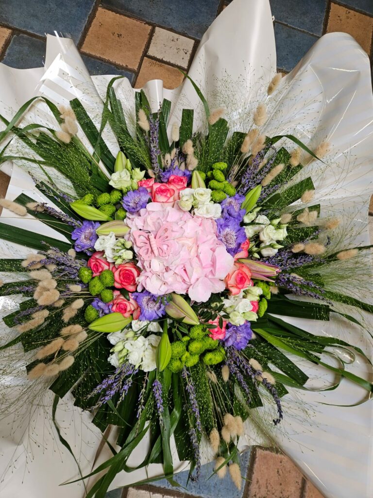 Composition ronde pastel : hortensia rose orné d'un papillon décoratif, roses fuchsia, scabieuses mauves, chrysanthèmes verts, lavande séchée et lagurus