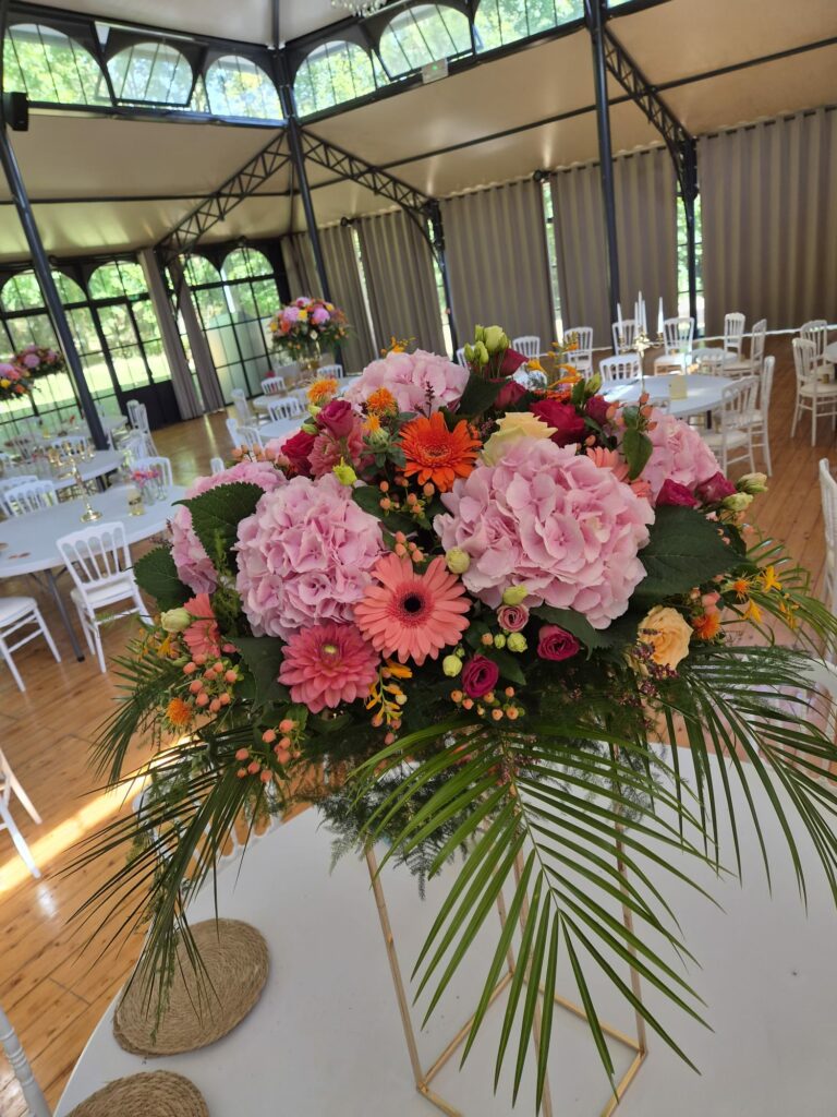Grande composition florale sur pied dans une orangerie dressée pour un mariage : hortensias roses, gerberas corail et orange, dahlias, roses et palmes