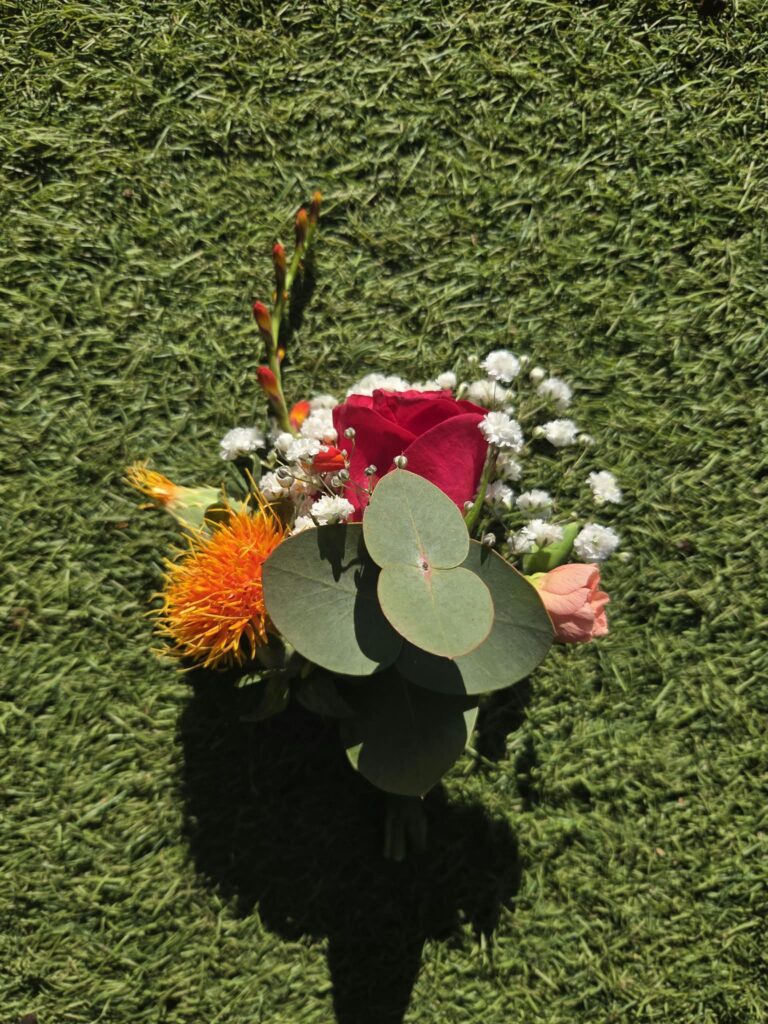 Boutonnière de mariage colorée : rose rouge, carthame orange, crocosmia, rose pêche, gypsophile et feuilles d'eucalyptus