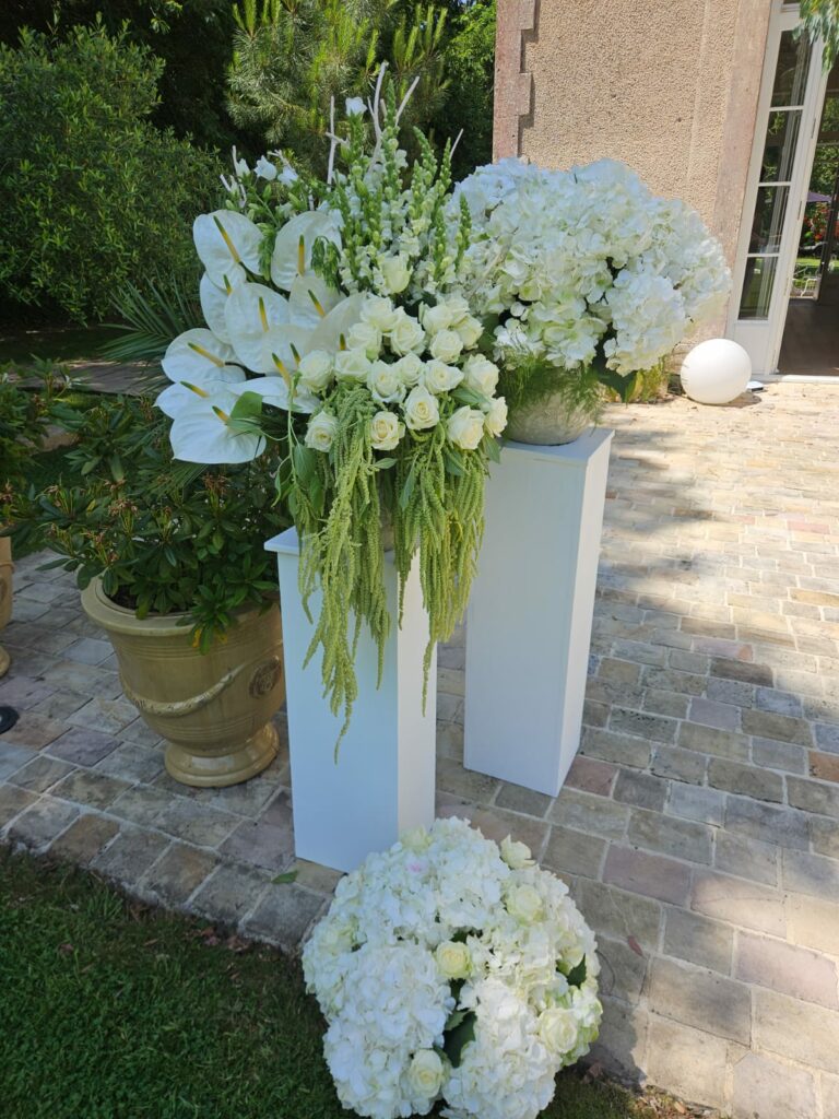 Compositions florales blanches posées sur des colonnes devant une bâtisse : anthuriums blancs, roses, hortensias, muflier et amarante verte retombante