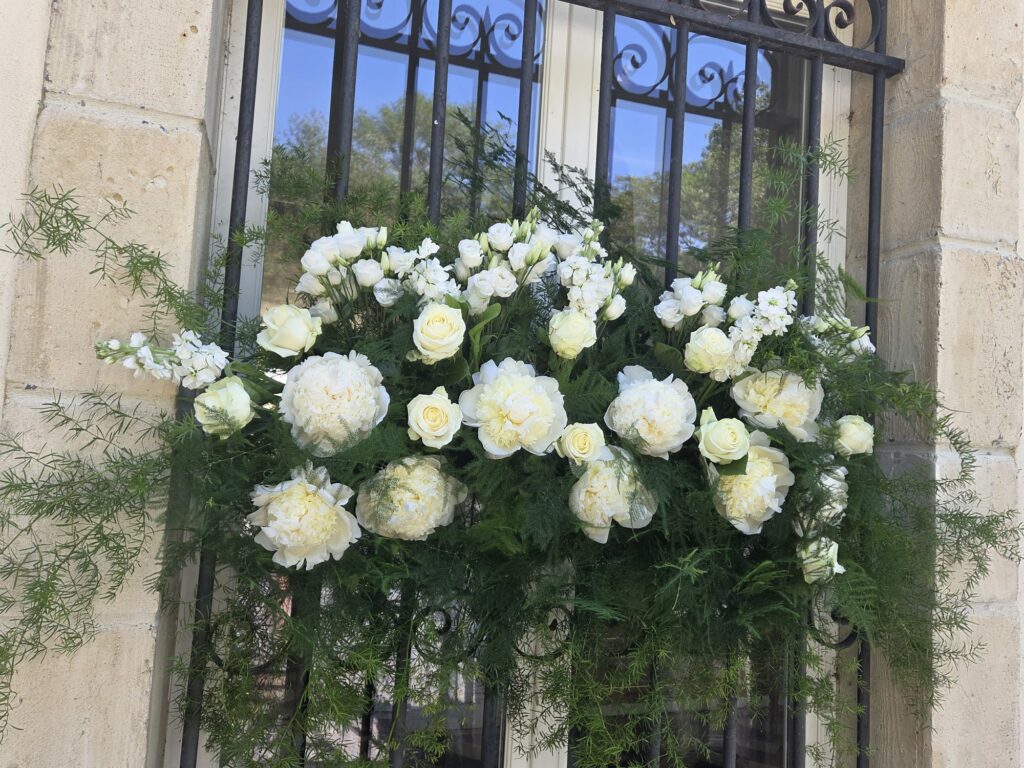 Composition florale blanche accrochée à une fenêtre à grille en fer forgé : pivoines, roses blanches, lisianthus, giroflées et feuillage d'asparagus retombant