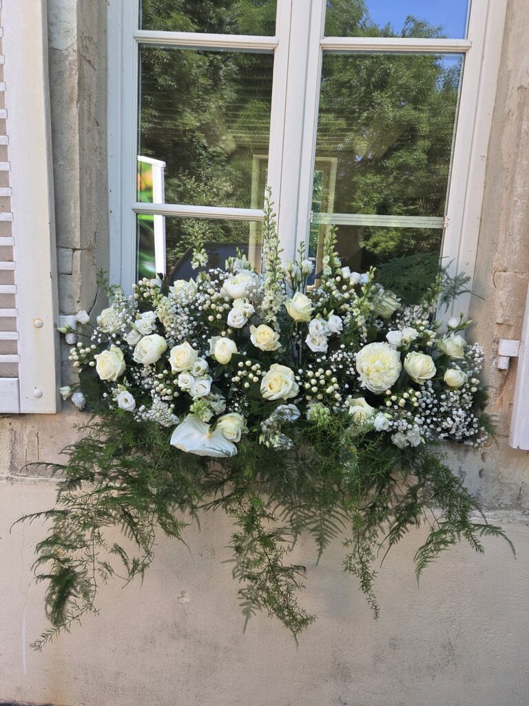 Composition florale entièrement blanche posée sur un appui de fenêtre en pierre : roses blanches, pivoines, lisianthus, gypsophile, anthurium et feuillage d'asparagus retombant