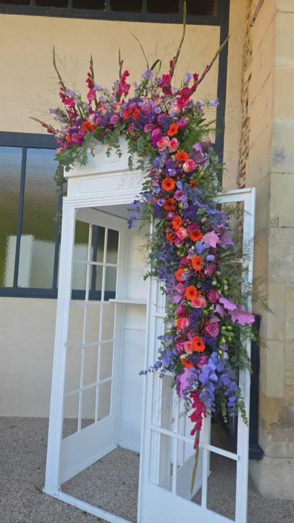 Décor floral en cascade sur une porte ancienne blanche : glaïeuls fuchsia, roses parme, gerberas orange, lisianthus violets et eucalyptus teinté
