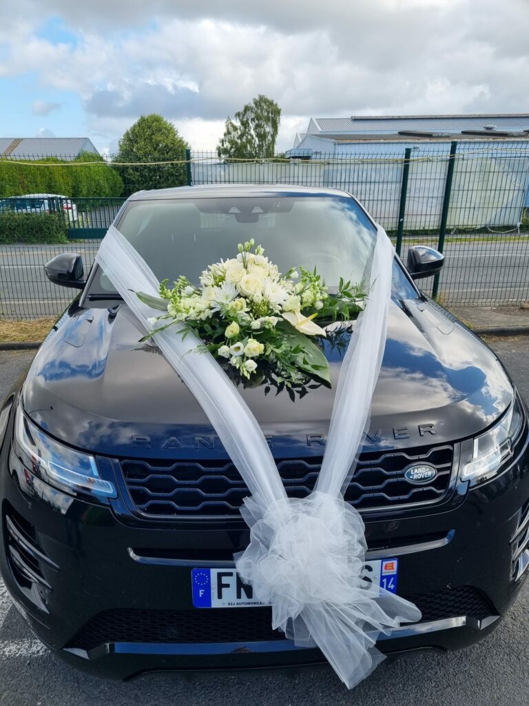 Décoration de voiture de mariage : composition de roses et lisianthus blancs posée sur le capot, avec des rubans de tulle blanc noués sur la calandre