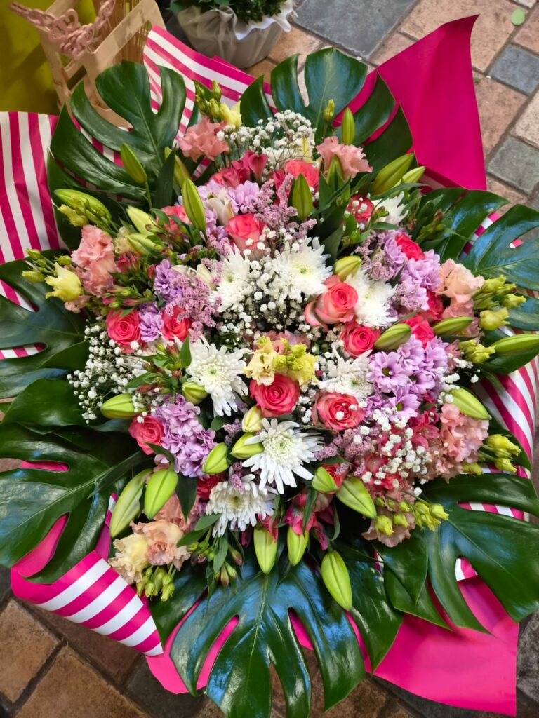 Grand bouquet rond aux tons roses et parme : roses fuchsia, jacinthes mauves, chrysanthèmes blancs, lisianthus, gypsophile et boutons de lis, sur de larges feuilles de monstera